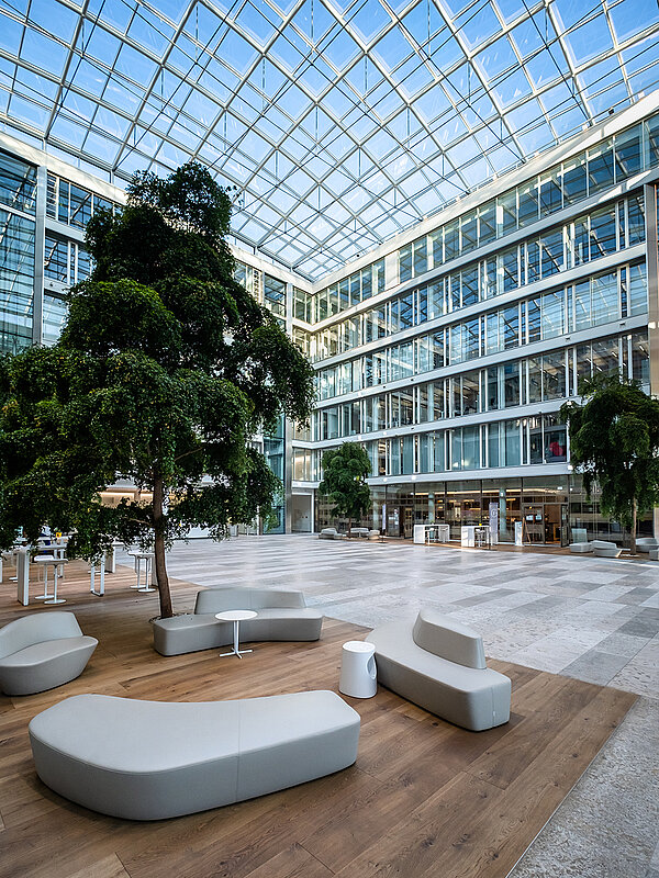 Modernes Atrium des Bürogebäudes Newton der TÜV Süd Gruppe in München mit Glasdach, großen Bäumen und stilvollen Sitzmöbeln auf Holzfußboden.