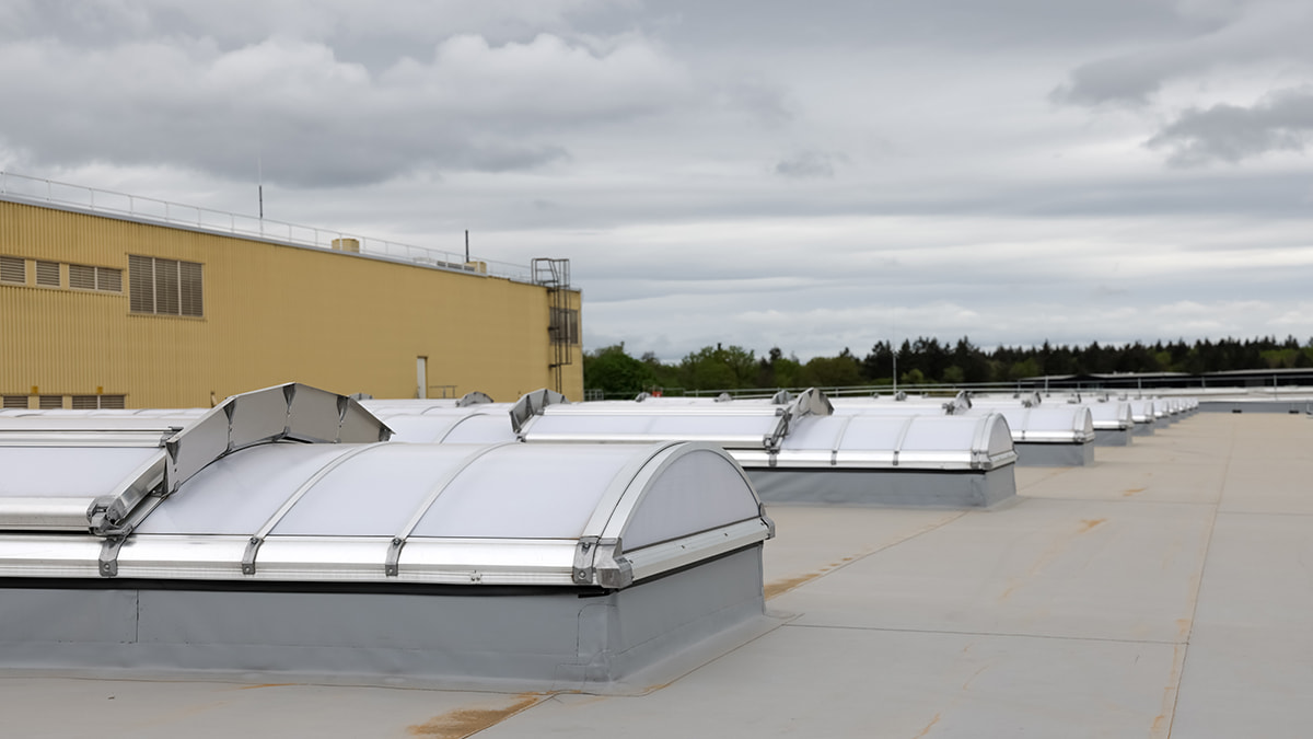 Mehrere neue Dachoberlichter auf einem großen Flachdach mit bewölktem Himmel im Hintergrund.